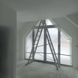 Grzegorz Wojdyła Gregor Bau - Pokój w trakcie prac wykończeniowych, z drabiną opartą o okno zabezpieczone folią malarską i świeżo zagruntowanymi ścianami.