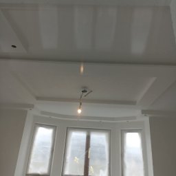 Grzegorz Wojdyła Gregor Bau - Pokój w trakcie wykańczania: świeżo wyszpachlowany sufit z dekoracyjnymi elementami, okna zabezpieczone folią malarską, widoczne narzędzia malarskie i oświetlenie budowlane.