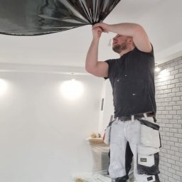 Grzegorz Wojdyła Gregor Bau - Pracownik na drabinie naciąga czarną folię na suficie w pomieszczeniu z białą ścianą i fragmentem ściany z cegły, widoczna skrzynka z narzędziami i zabezpieczone meble.