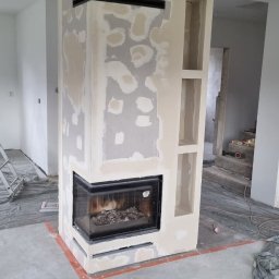 Grzegorz Wojdyła Gregor Bau - Wnętrze w trakcie remontu: obudowa kominka z wkładem, częściowo wyszpachlowana, z czarnymi dekoracyjnymi elementami poziomymi i wnękami, podłoga zabezpieczona folią, widoczne ślady szpachlowania.