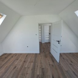 Grzegorz Wojdyła Gregor Bau - Poddasze po remoncie z drewnianą podłogą, białe ściany, otwarte białe drzwi z czarną klamką i widok na kolejne pomieszczenie z identycznymi drzwiami.