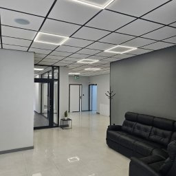 Grzegorz Wojdyła Gregor Bau - Nowoczesne biuro z podwieszanym sufitem kasetonowym z oświetleniem LED, czarną skórzaną sofą i błyszczącą podłogą z płytek ceramicznych.