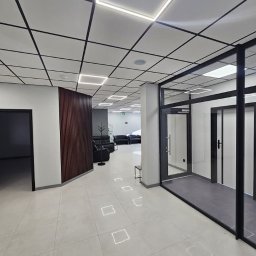 Grzegorz Wojdyła Gregor Bau - Nowoczesne wnętrze biurowe z podwieszanym sufitem kasetonowym z oświetleniem LED, szklane ścianki działowe z czarnymi ramami i płytki podłogowe z wbudowanymi punktami świetlnymi.