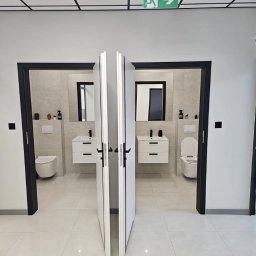 Grzegorz Wojdyła Gregor Bau - Nowoczesna łazienka w biurze z dwoma kabinami, widok przez częściowo otwarte składane drzwi, białe płytki podłogowe, czarne ramy drzwiowe i białe umywalki z szafkami.