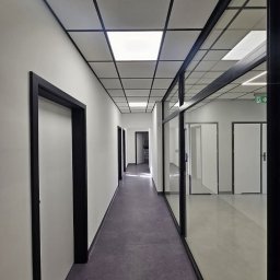 Grzegorz Wojdyła Gregor Bau - Korytarz biurowy z białymi ścianami, ciemnymi ościeżnicami drzwi i szarą podłogą, widok w głąb z przeszkloną ścianą po prawej stronie.