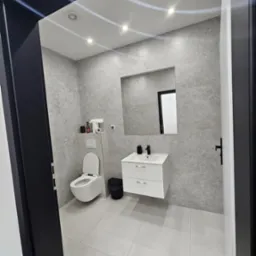 Nowoczesna łazienka z szarymi ścianami imitującymi beton, białą wiszącą toaletą i umywalką z szafką, widoczna przez czarną futrynę drzwi.