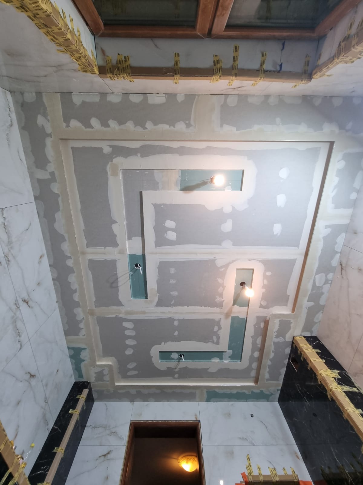 Sufit podwieszany z płyt kartonowo-gipsowych w trakcie montażu w łazience, widoczne profile, szpachlowanie łączeń i otwory na oświetlenie punktowe. Na ścianach białe płytki imitujące marmur.