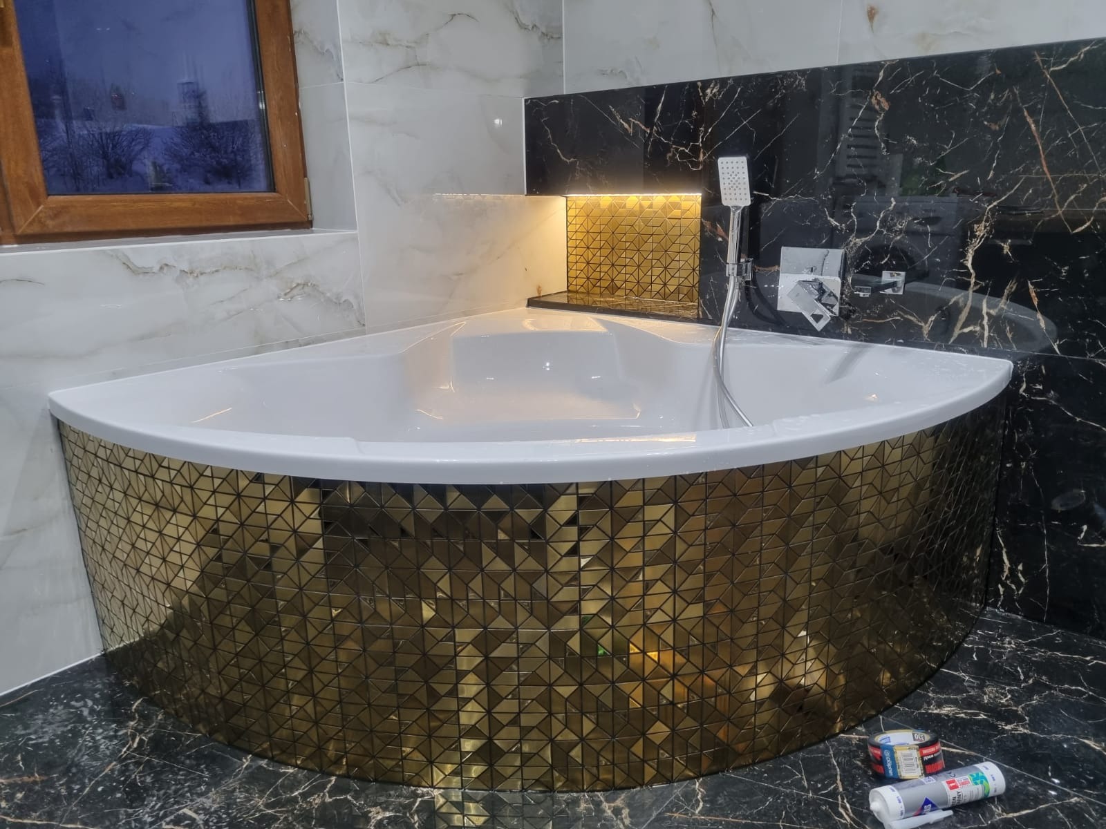 Narożna wanna obudowana złotą mozaiką, czarne marmurowe ściany i podłoga, widoczne akcesoria montażowe i okno w tle.