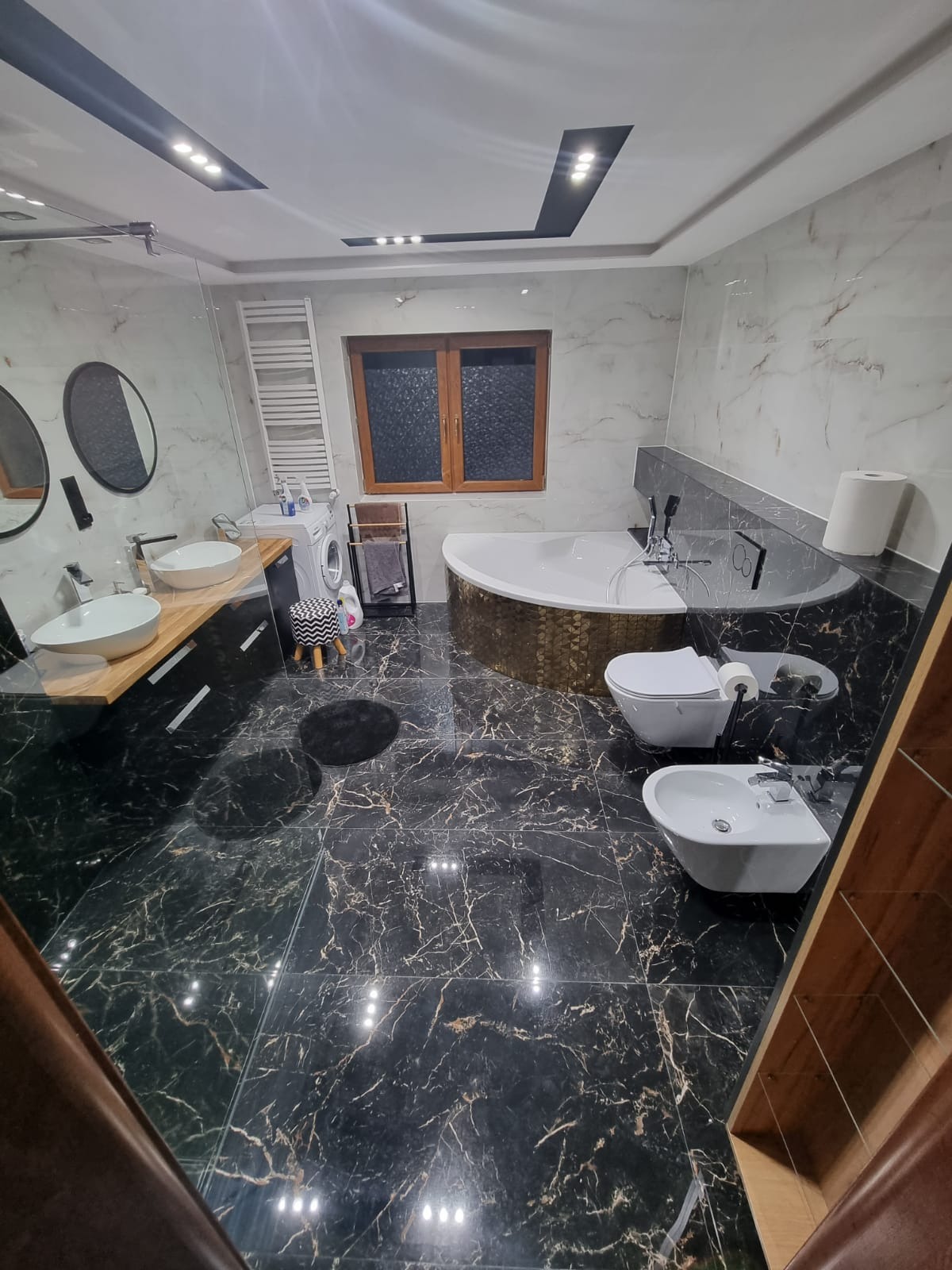 Elegancka łazienka z czarną marmurową podłogą i ścianami w odcieniach bieli, wyposażona w owalną wannę z mozaikową obudową, wiszącą toaletę i bidet, umywalki nablatowe na drewnianym blacie, pralkę...