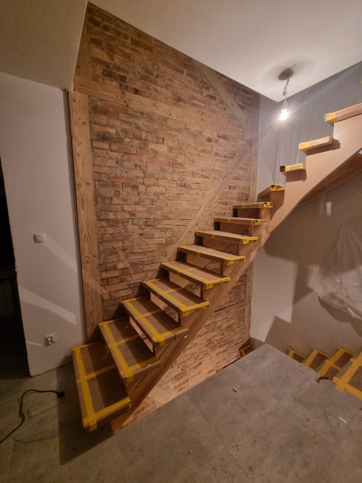 Drewniane schody z zabezpieczeniami z żółtej taśmy malarskiej, zamontowane przy ścianie z czerwonej cegły i drewnianej ramy, widok z dołu na górę.