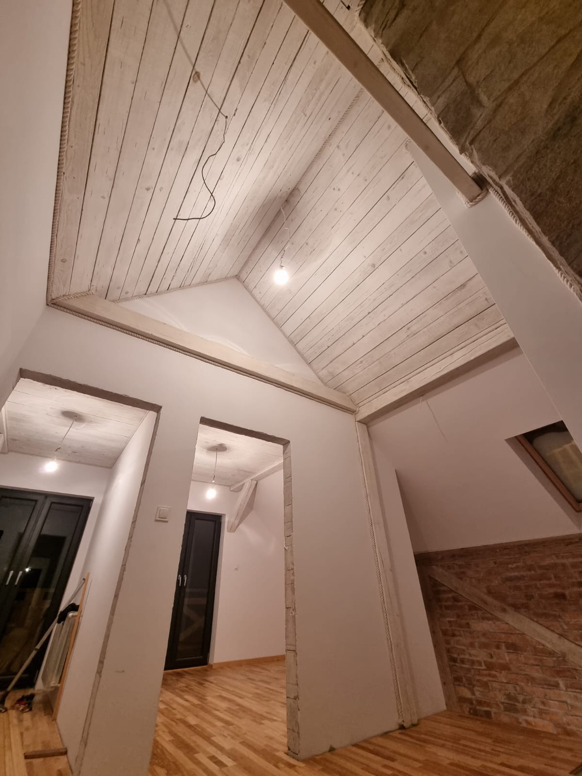 Poddasze z drewnianym, pomalowanym na biało sufitem, widoczne przygotowane otwory drzwiowe z surowymi ościeżnicami i wiszące kable od oświetlenia.