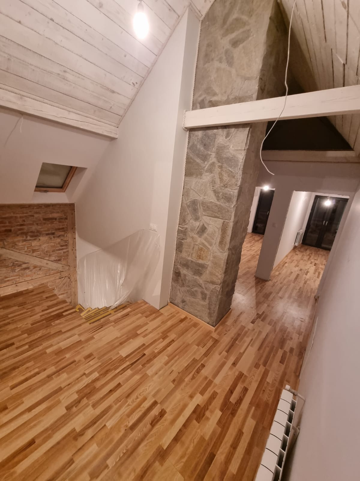 Wnętrze domu w trakcie remontu z drewnianą podłogą, kolumną obłożoną kamieniem, widoczną ścianą z cegły i zabezpieczoną folią, oraz fragmentem grzejnika.