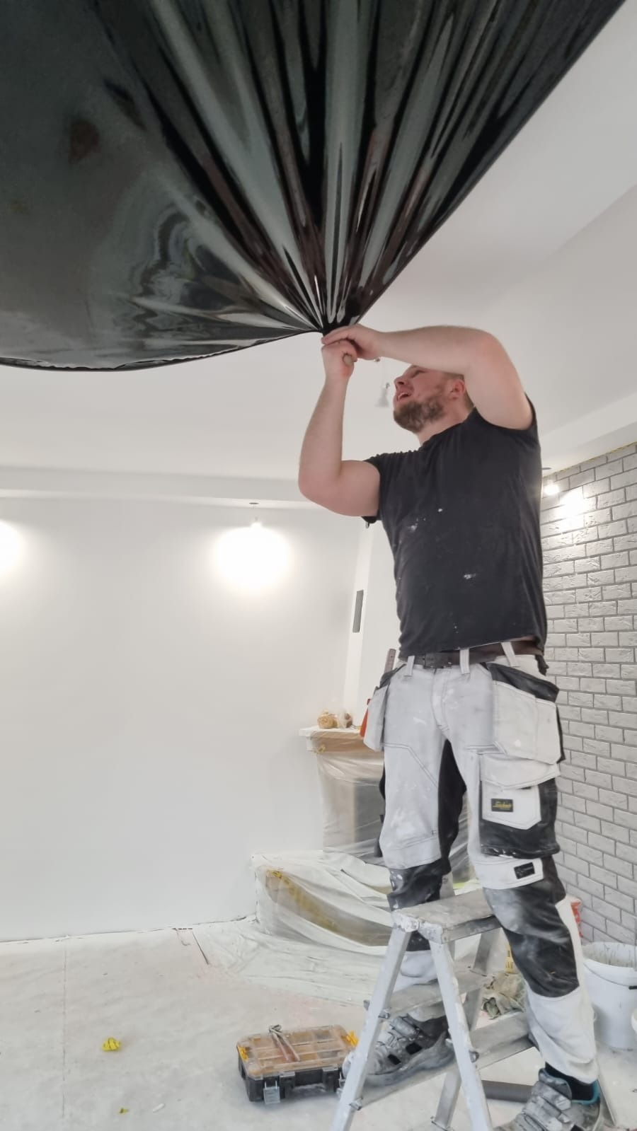 Pracownik na drabinie montuje błyszczący, czarny sufit napinany w pomieszczeniu z białą ścianą i fragmentem ściany wykończonej imitacją cegły.