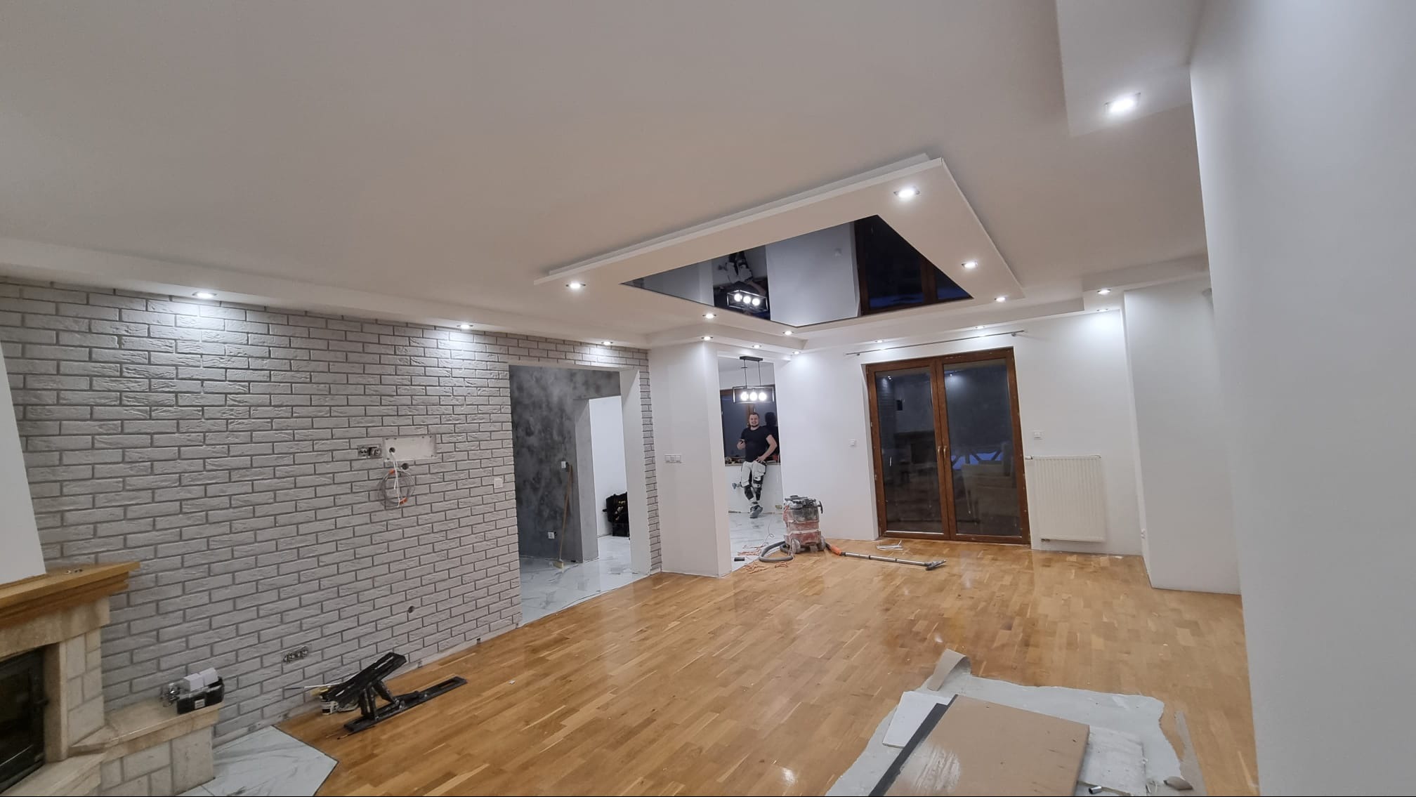 Wnętrze w trakcie prac wykończeniowych: ściana wyłożona cegłą dekoracyjną, drewniana podłoga, sufit z podwieszaną, lustrzaną częścią, widoczne narzędzia i osoba w tle.