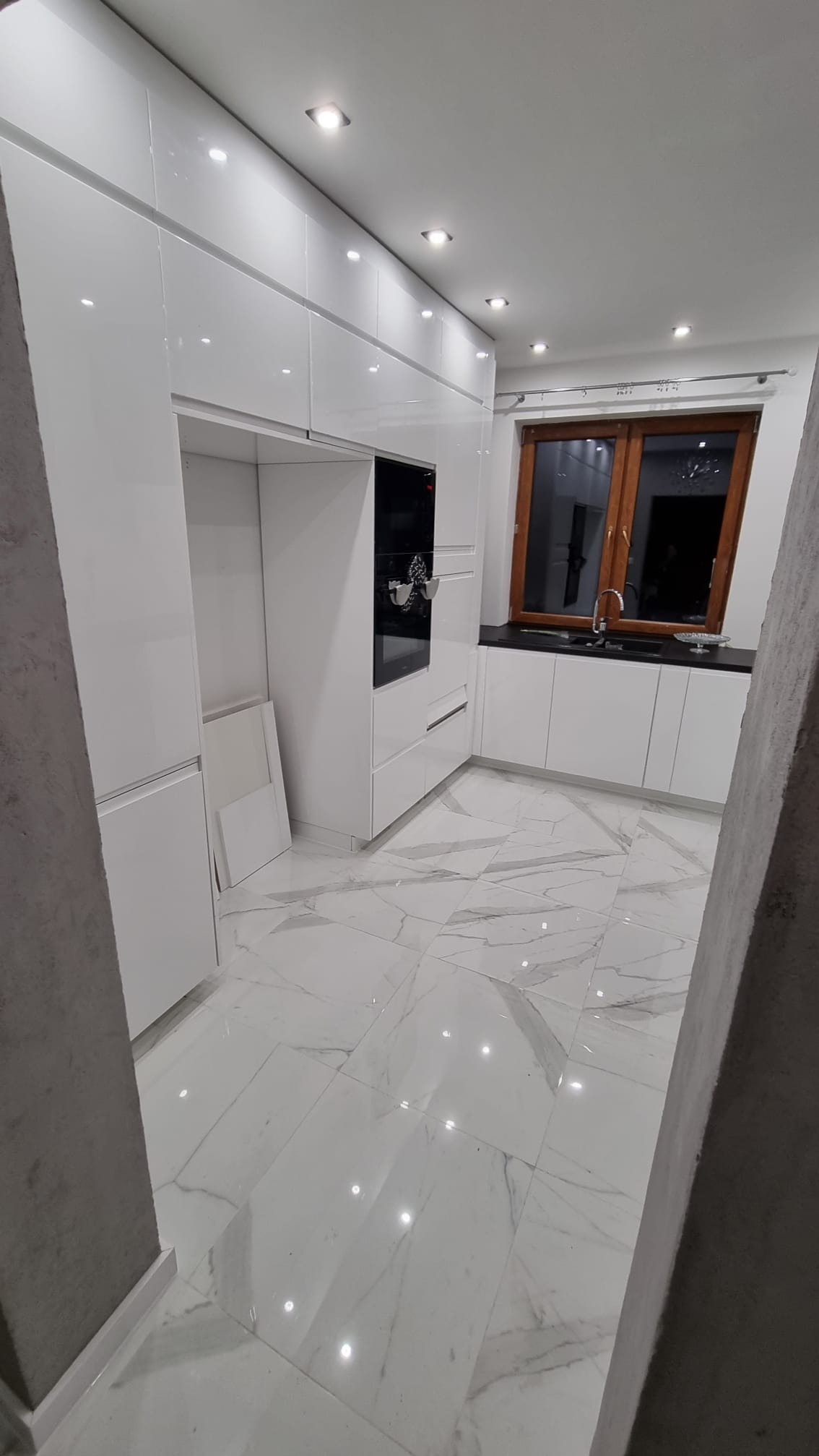 Nowoczesna, biała kuchnia z połyskiem w Łobozew Górny, z marmurową posadzką i oknem w drewnianej ramie. Widoczne oświetlenie sufitowe i wbudowany piekarnik.