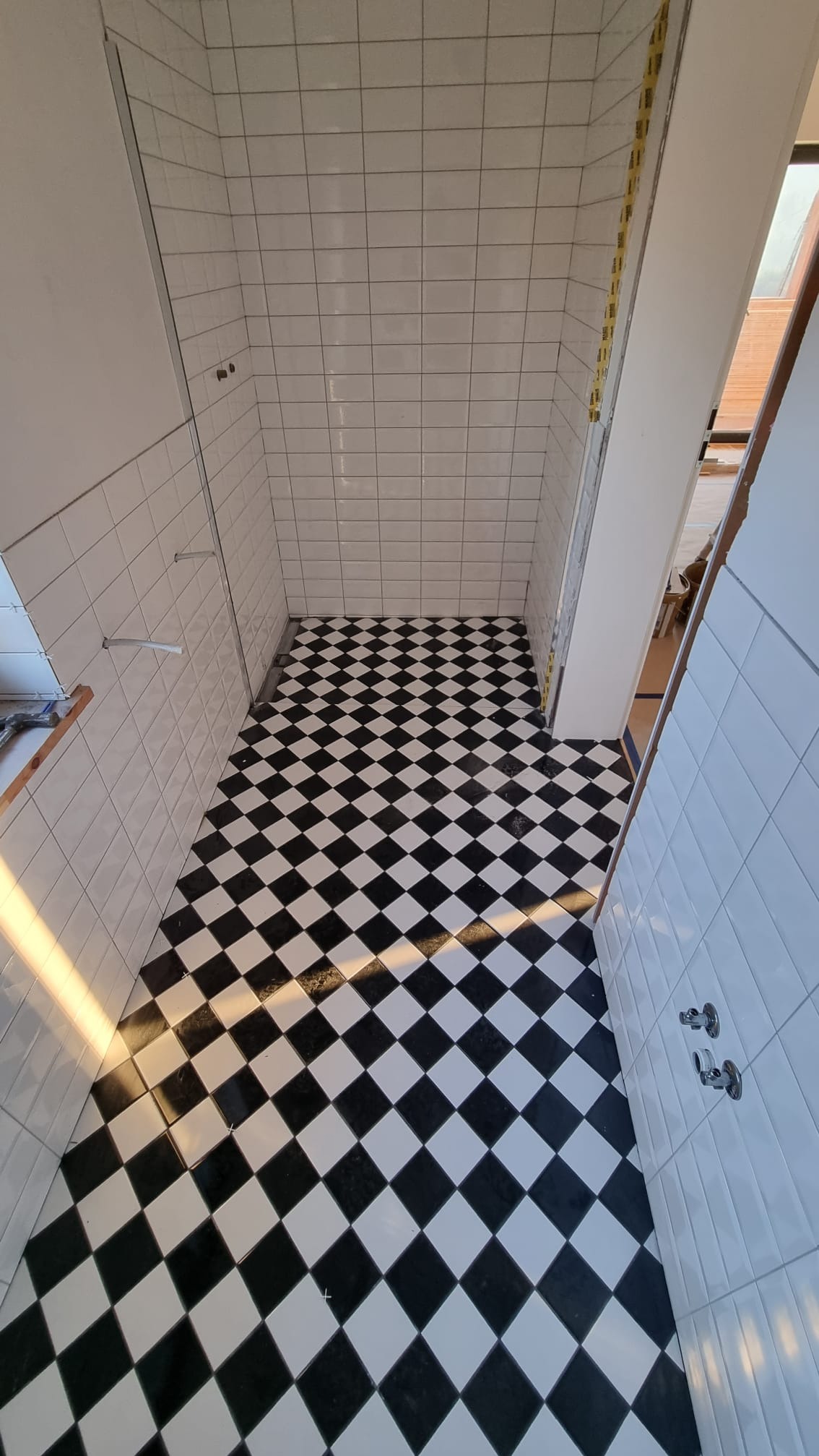 Wnętrze łazienki w trakcie wykańczania, z ułożoną czarno-białą szachownicą na podłodze i białymi płytkami na ścianach, widoczne niedokończone prace przy kabinie prysznicowej.