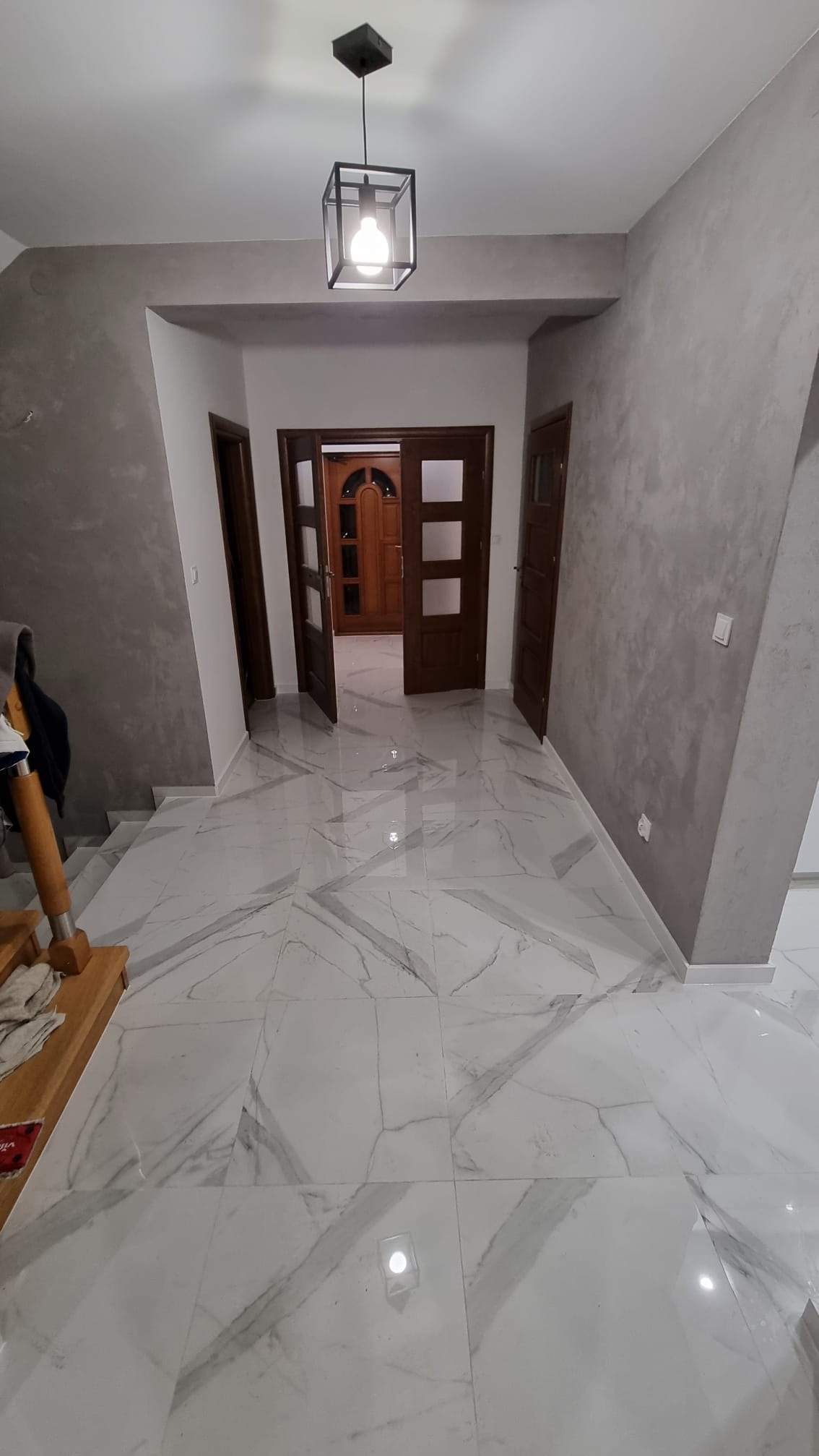 Wykończony korytarz z białymi marmurowymi płytkami podłogowymi, szarymi ścianami z efektem dekoracyjnym i ciemnymi drewnianymi drzwiami.