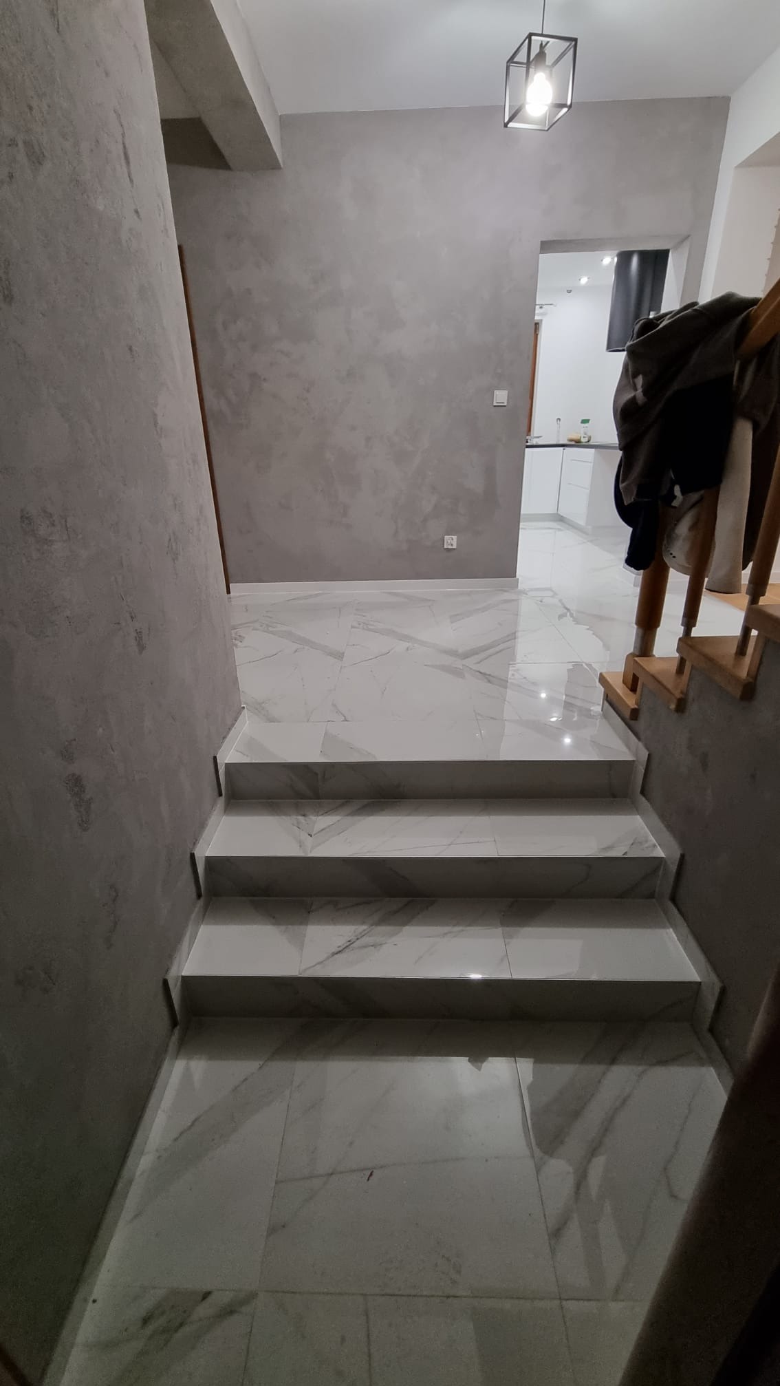 Wykończone schody z białego marmuru z szarymi żyłami prowadzące do przedpokoju z gładzią na ścianach i widokiem na białą kuchnię w tle.