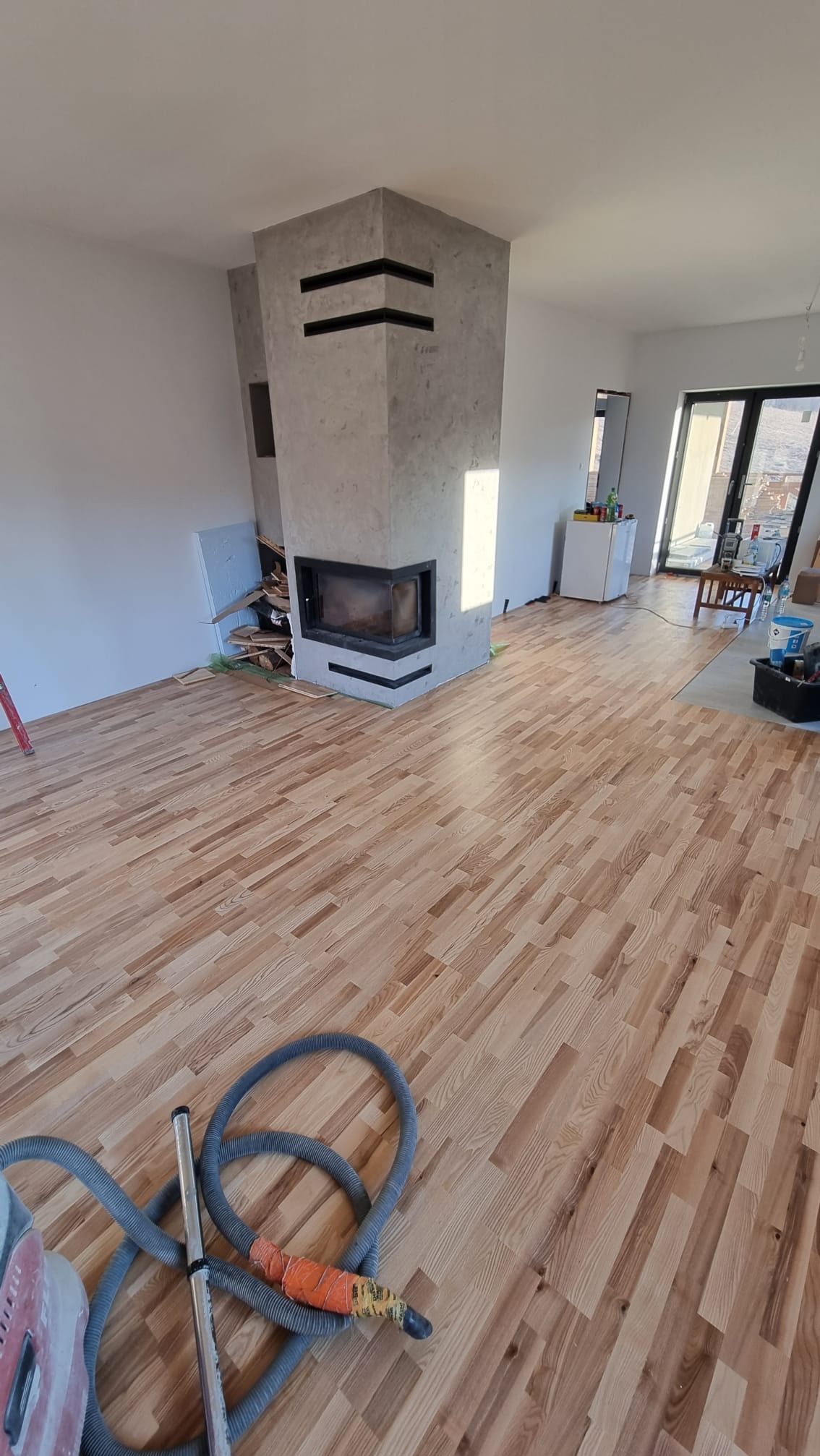 Nowa drewniana podłoga ułożona w jodełkę, widoczny kominek z szarą obudową i czarnymi akcentami, częściowo wykończone ściany, odkurzacz przemysłowy z wężem na podłodze.