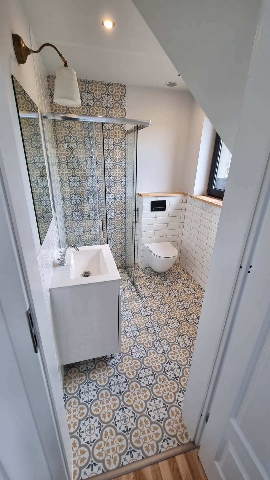 Wykończona łazienka w skosach z kabiną prysznicową, umywalką z szafką, toaletą podwieszaną i wzorzystą, kolorową podłogą oraz częściowo wykończona białą glazurą.