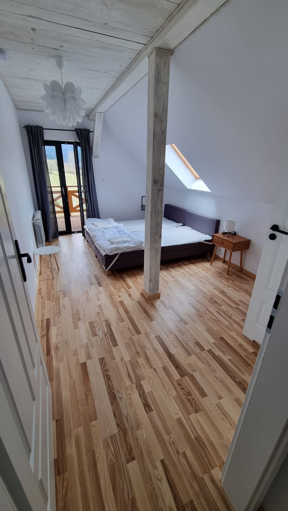 Jasne wnętrze pokoju na poddaszu z drewnianą podłogą, łóżkiem z fioletowym zagłówkiem, drewnianą kolumną i wyjściem na balkon z widokiem na zieleń.