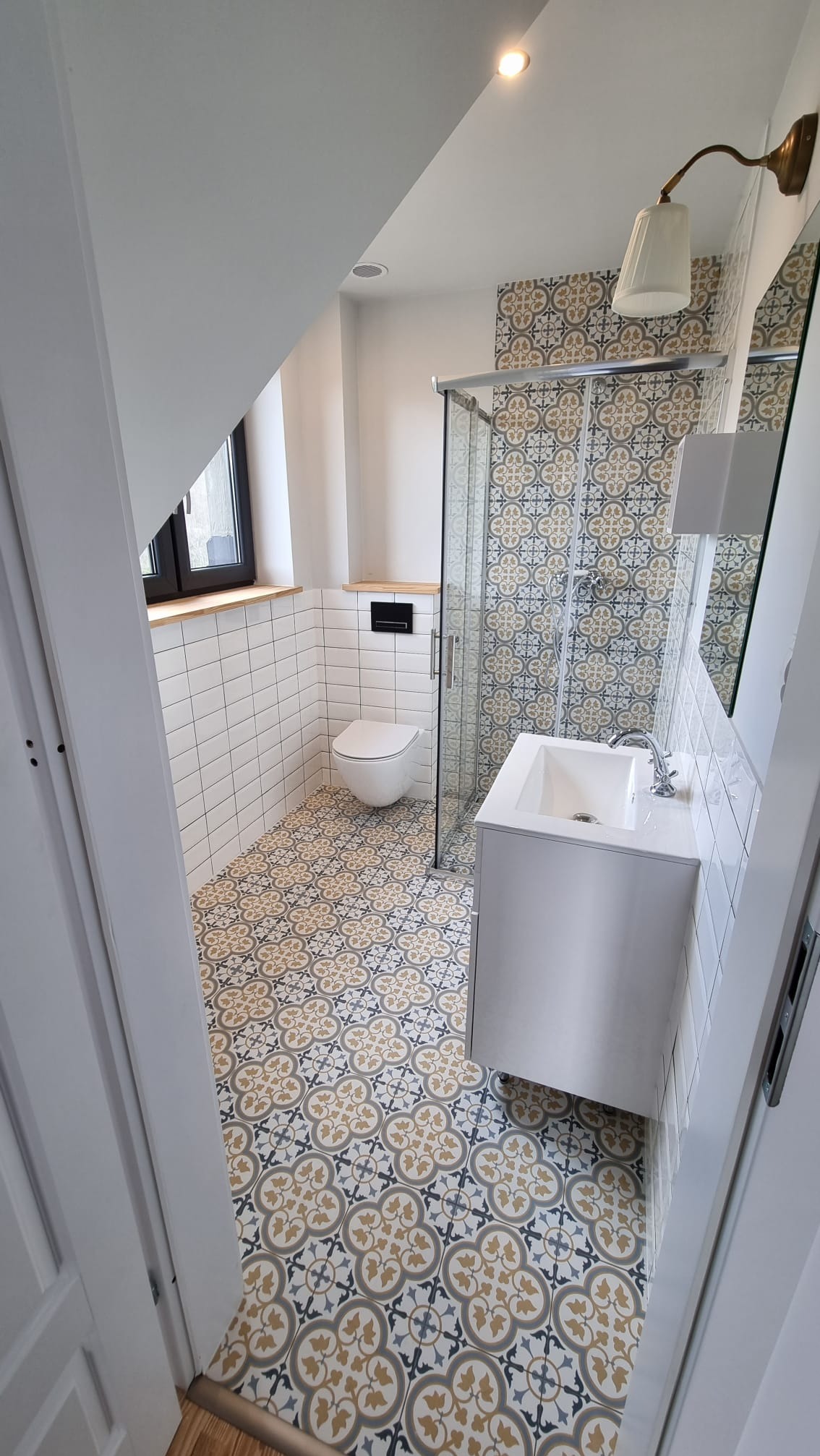 Ukończona łazienka w stylu retro z wzorzystą podłogą i ścianą pod prysznicem, białe płytki na ścianach, wisząca toaleta i umywalka, oświetlenie punktowe i kinkiet w stylu vintage.