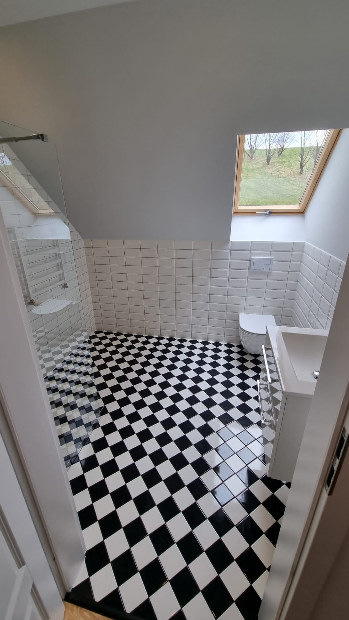 Wnętrze wyremontowanej łazienki na poddaszu z czarno-białą szachownicą na podłodze, białe kafelki na ścianach, kabina prysznicowa ze szklaną ścianką, umywalka z szafką i toaleta w zabudowie, okno...