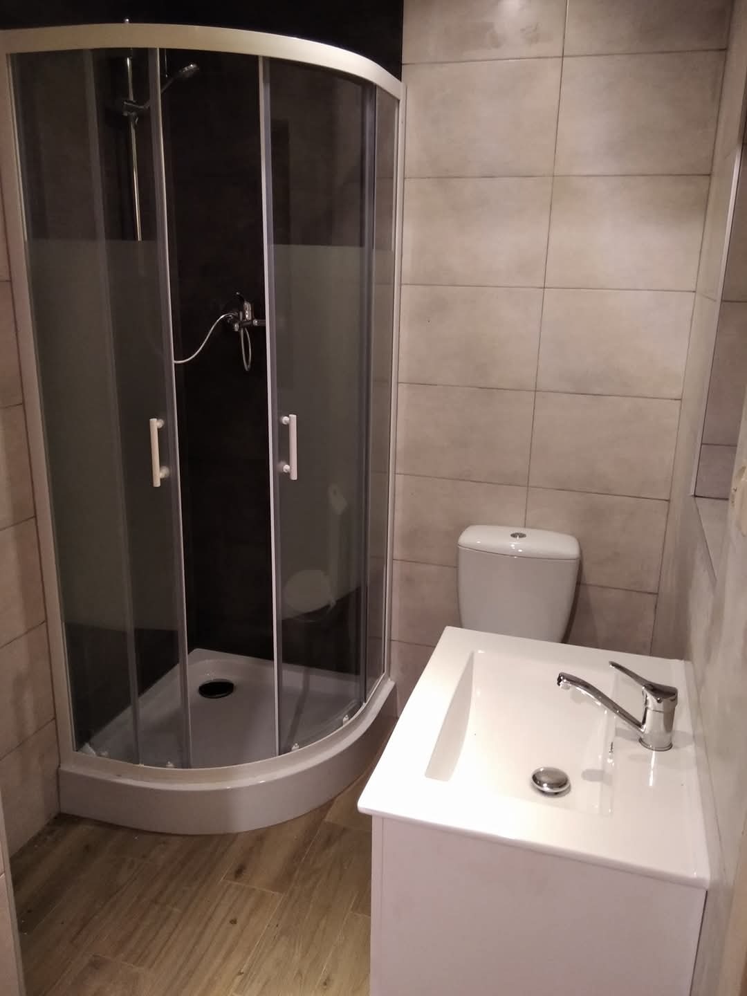 Wyremontowana łazienka z kabiną prysznicową z przesuwnymi drzwiami, białą umywalką z szafką i toaletą w tle, wykończona jasnymi płytkami.