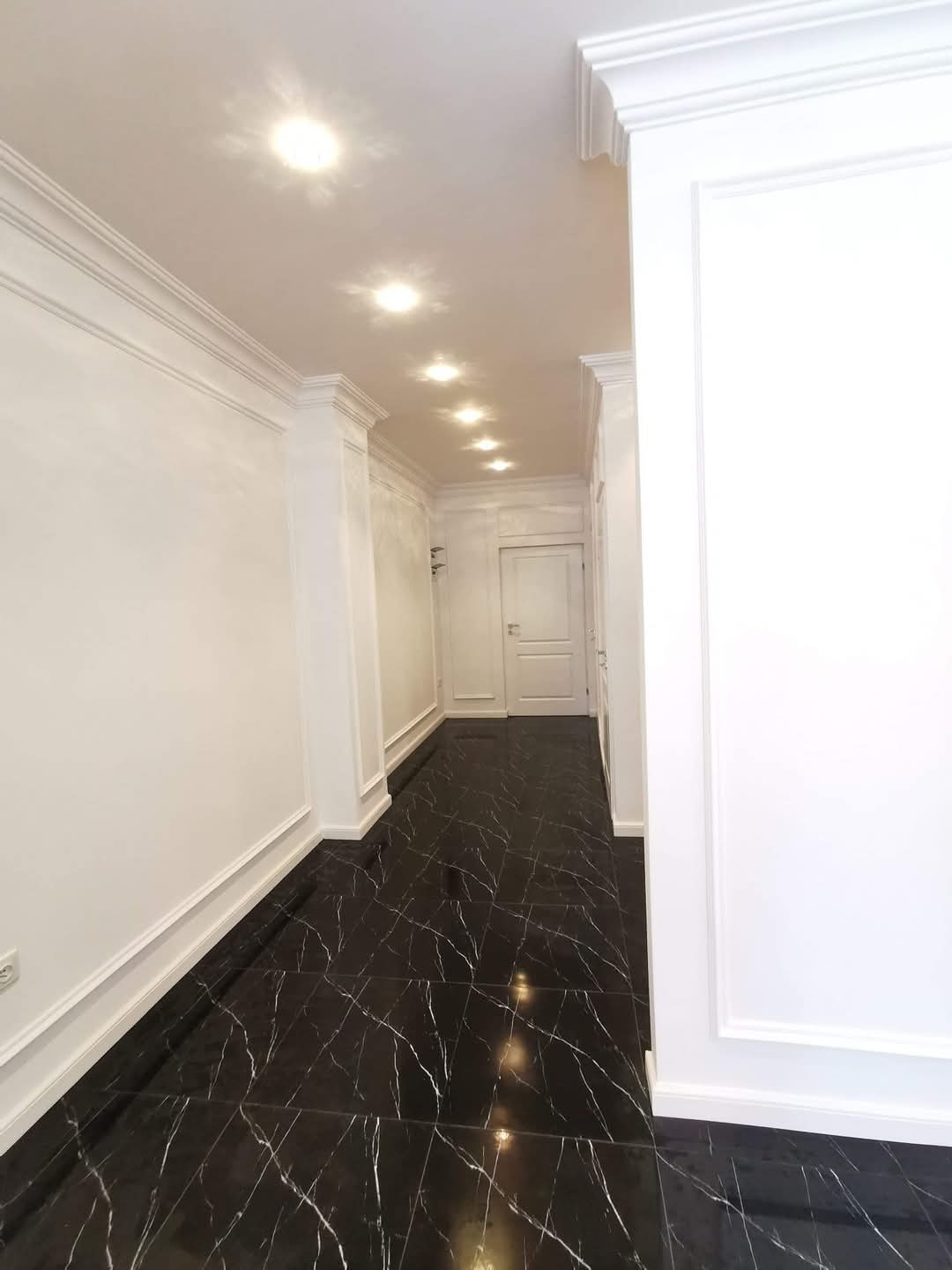 Wykończony korytarz z czarną marmurową podłogą, białymi ścianami z dekoracyjnymi listwami i rzędem punktowych lamp sufitowych.