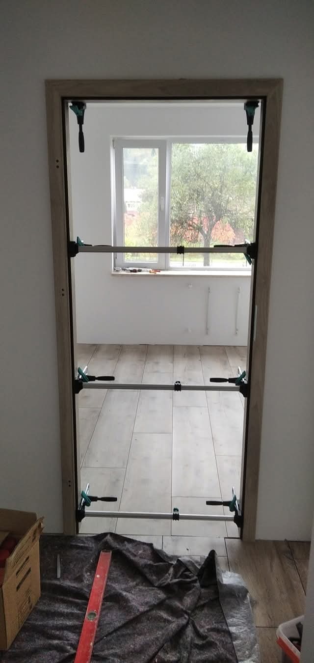 Ościeżnica drzwiowa mocowana rozpórkami teleskopowymi w nowym otworze, widoczny poziomica, karton i zabezpieczenie podłogi.