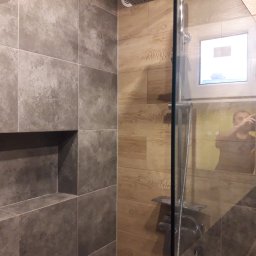 P.REM-BUD PRZEMYSLAW PIOTROWSKI USŁUGI REMONTOWO-BUDOWLANE - Nowoczesna kabina prysznicowa z szarymi płytkami imitującymi kamień i drewnopodobnymi płytkami na ścianie, widoczny wnęka w ścianie wyłożona szarymi płytkami, szklane drzwi kabiny, w odbiciu osoba...