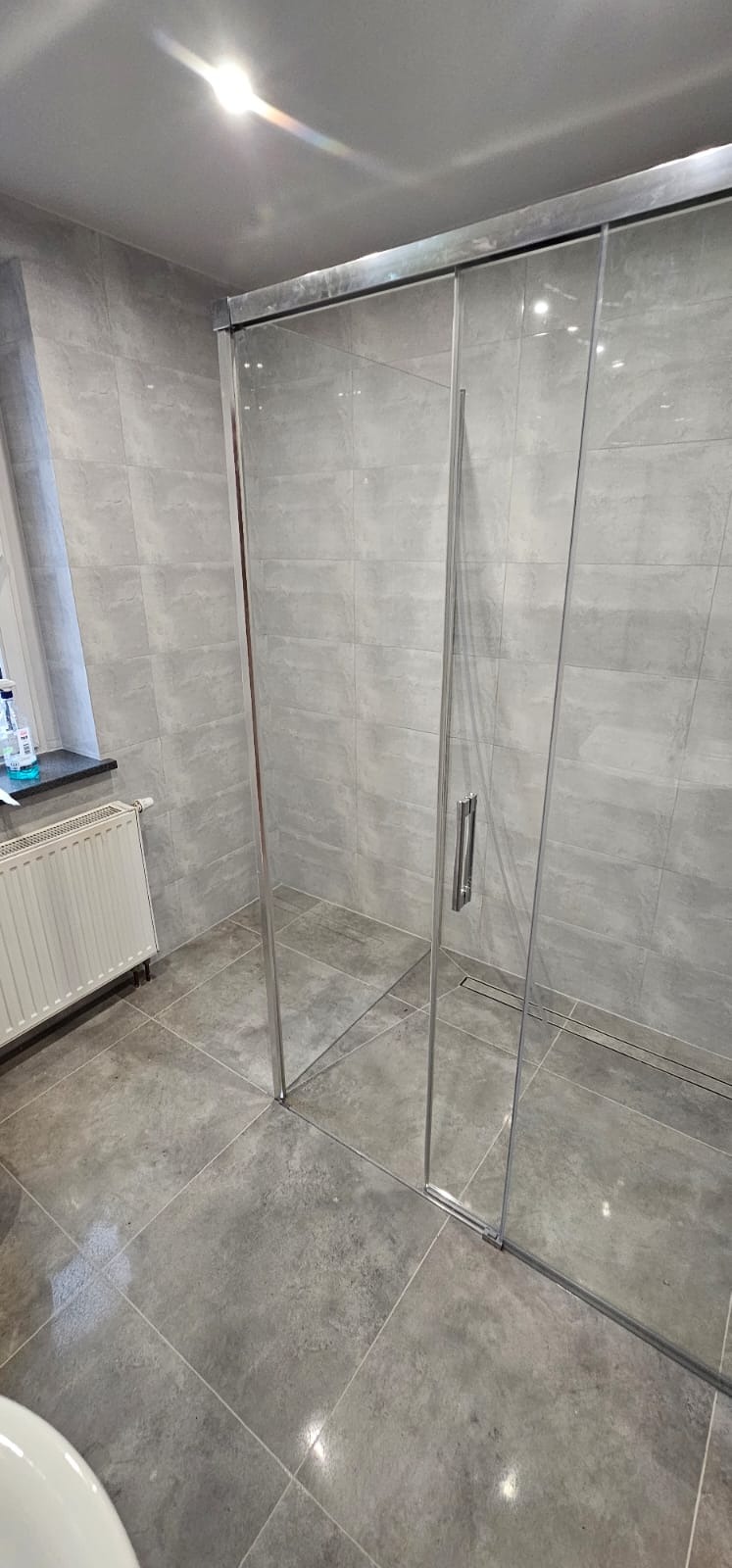 Nowoczesna kabina prysznicowa z przesuwnymi drzwiami, szare płytki na ścianach i podłodze, minimalistyczny odpływ liniowy w podłodze, widoczny fragment grzejnika i okna.