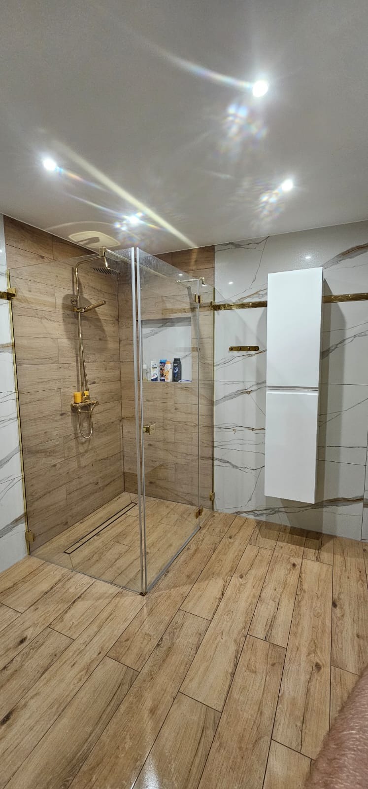 Nowoczesna kabina prysznicowa walk-in z odpływem liniowym, wykończona płytkami imitującymi drewno, z złotymi akcentami i białymi szafkami wiszącymi.