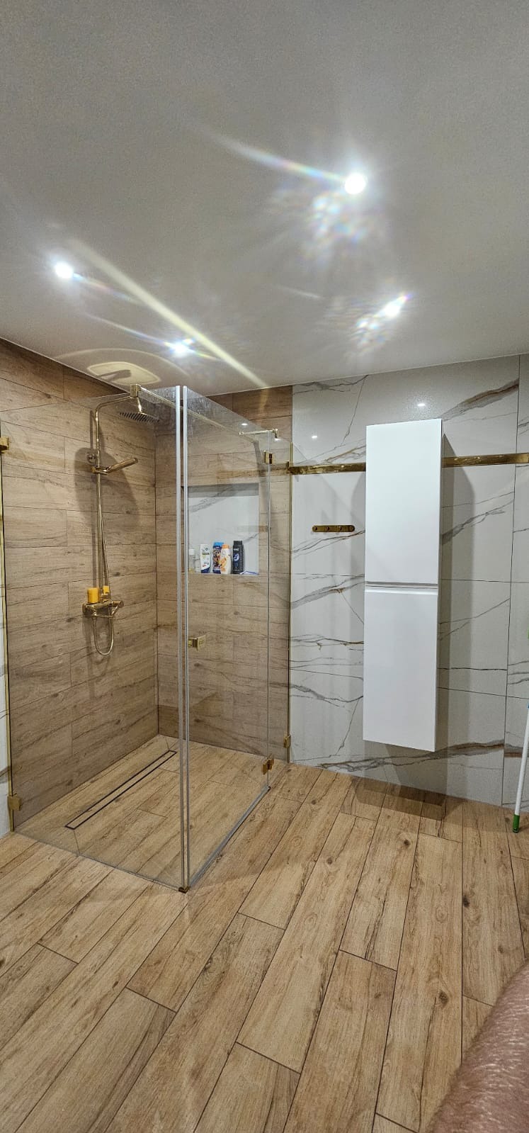 Nowoczesna kabina prysznicowa ze złotymi elementami, drewnopodobne płytki na podłodze i ścianach, wisząca biała szafka, widoczne kosmetyki w półce prysznicowej.