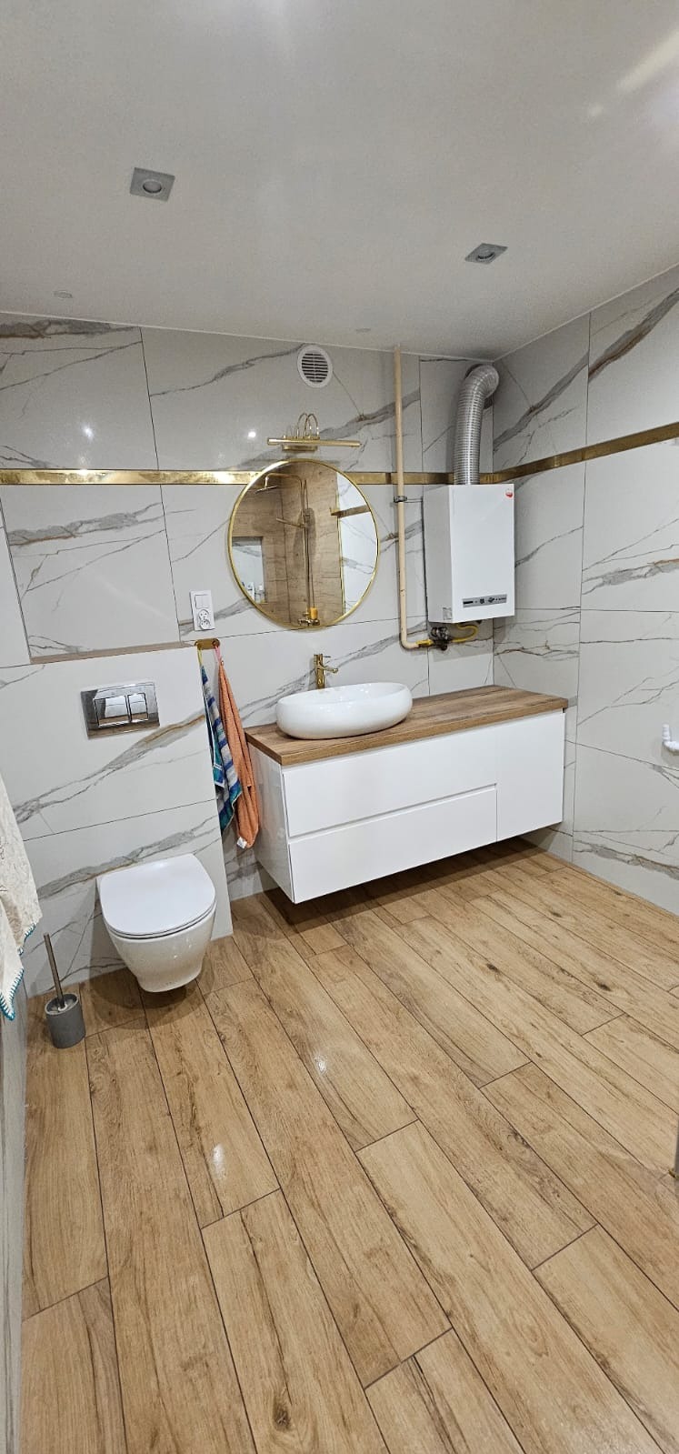 Wykończona łazienka w Lubomi z płytkami imitującymi marmur z złotymi akcentami, drewnopodobną podłogą, okrągłym lustrem i wiszącą szafką z umywalką nablatową.