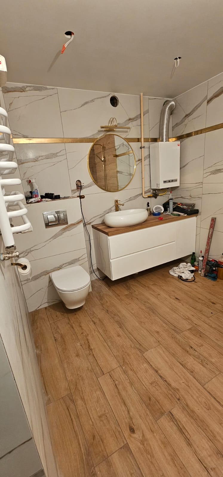 Nowoczesna łazienka w trakcie wykańczania, z marmurowymi płytkami, okrągłym lustrem w złotej ramie, białym umywalką nablatową, grzejnikiem drabinkowym i drewnopodobną podłogą; widoczne...