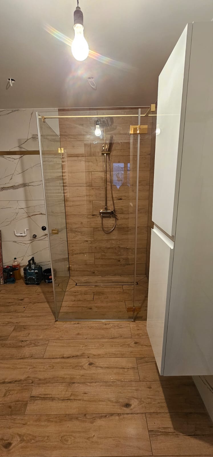 Kabina prysznicowa ze złotymi okuciami i drewnopodobnymi płytkami, widoczne narzędzia na podłodze, surowa żarówka.