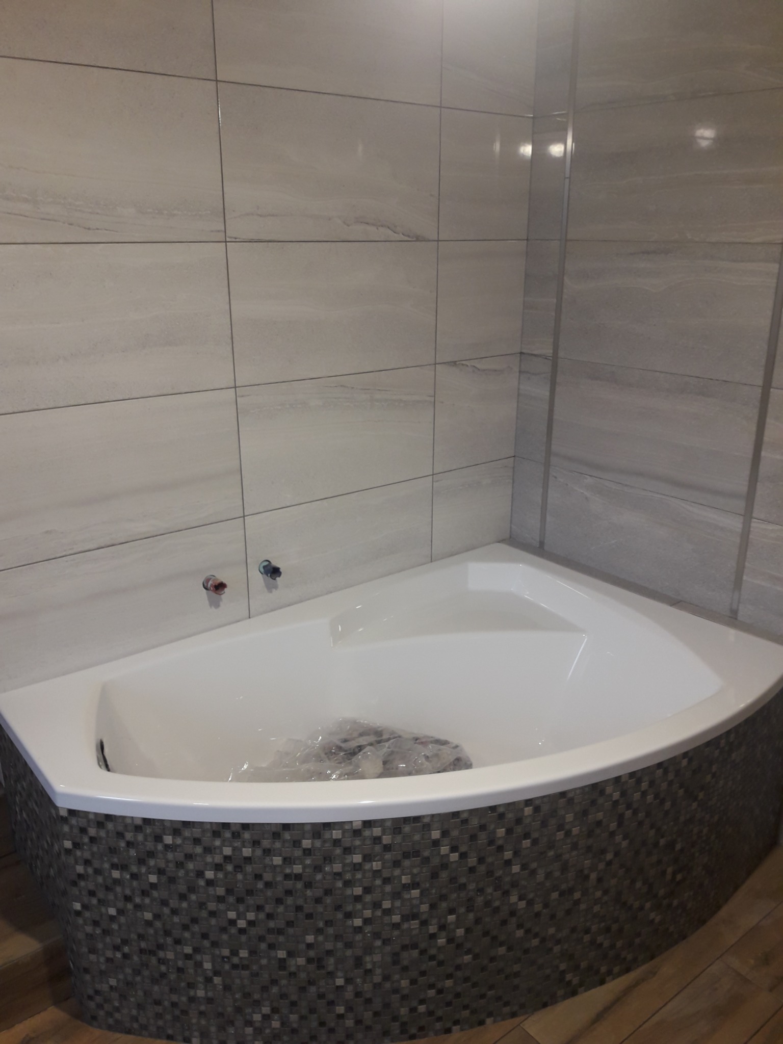Nowoczesna, narożna wanna obudowana mozaiką w odcieniach szarości, z jasnymi płytkami imitującymi marmur na ścianach i wystającymi zaworami. W wannie leżą folie ochronne.