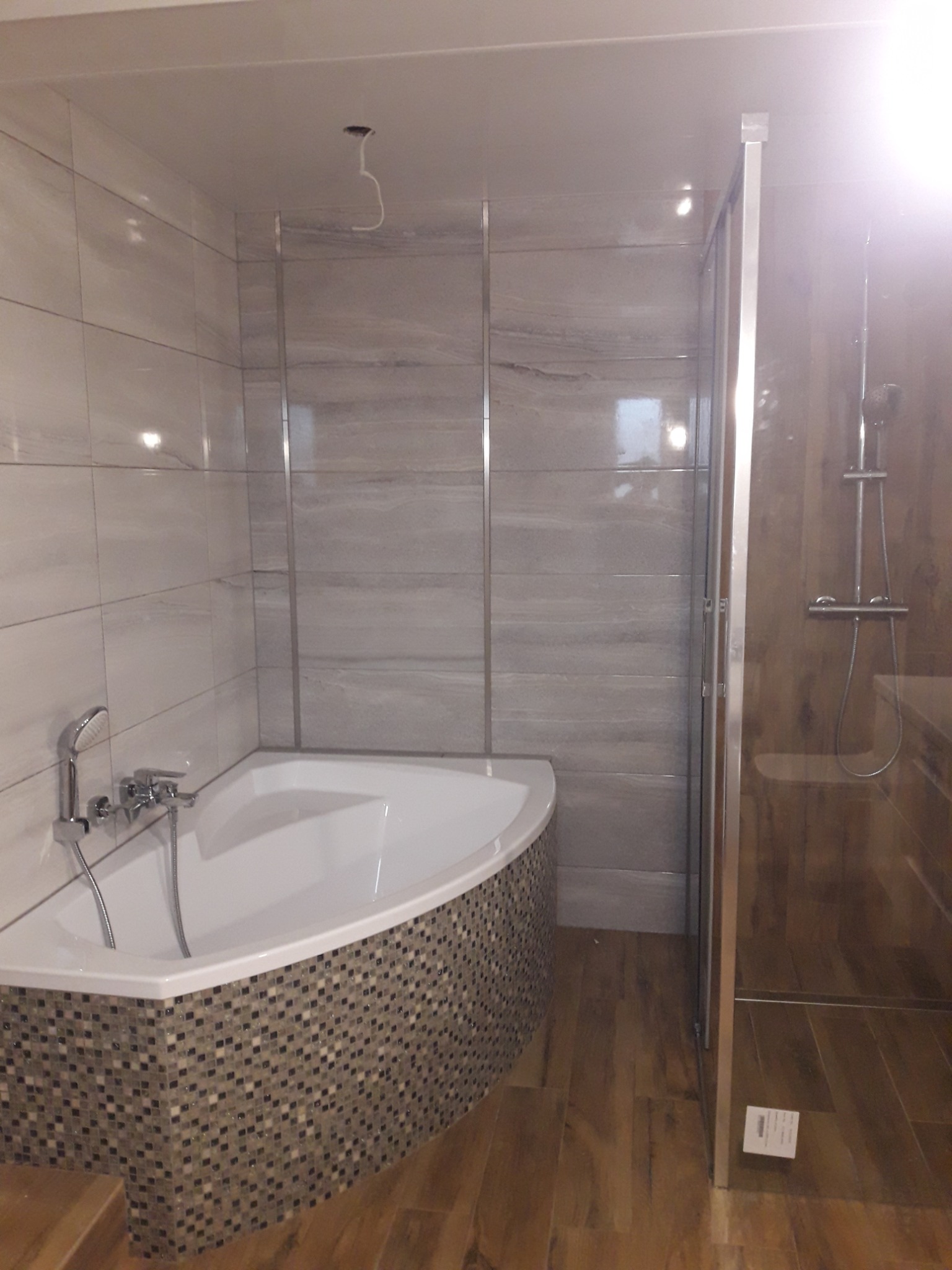 Nowoczesna łazienka z narożną wanną obłożoną mozaiką, szklaną kabiną prysznicową i płytkami imitującymi kamień na ścianach, widoczne niedokończone oświetlenie sufitowe.