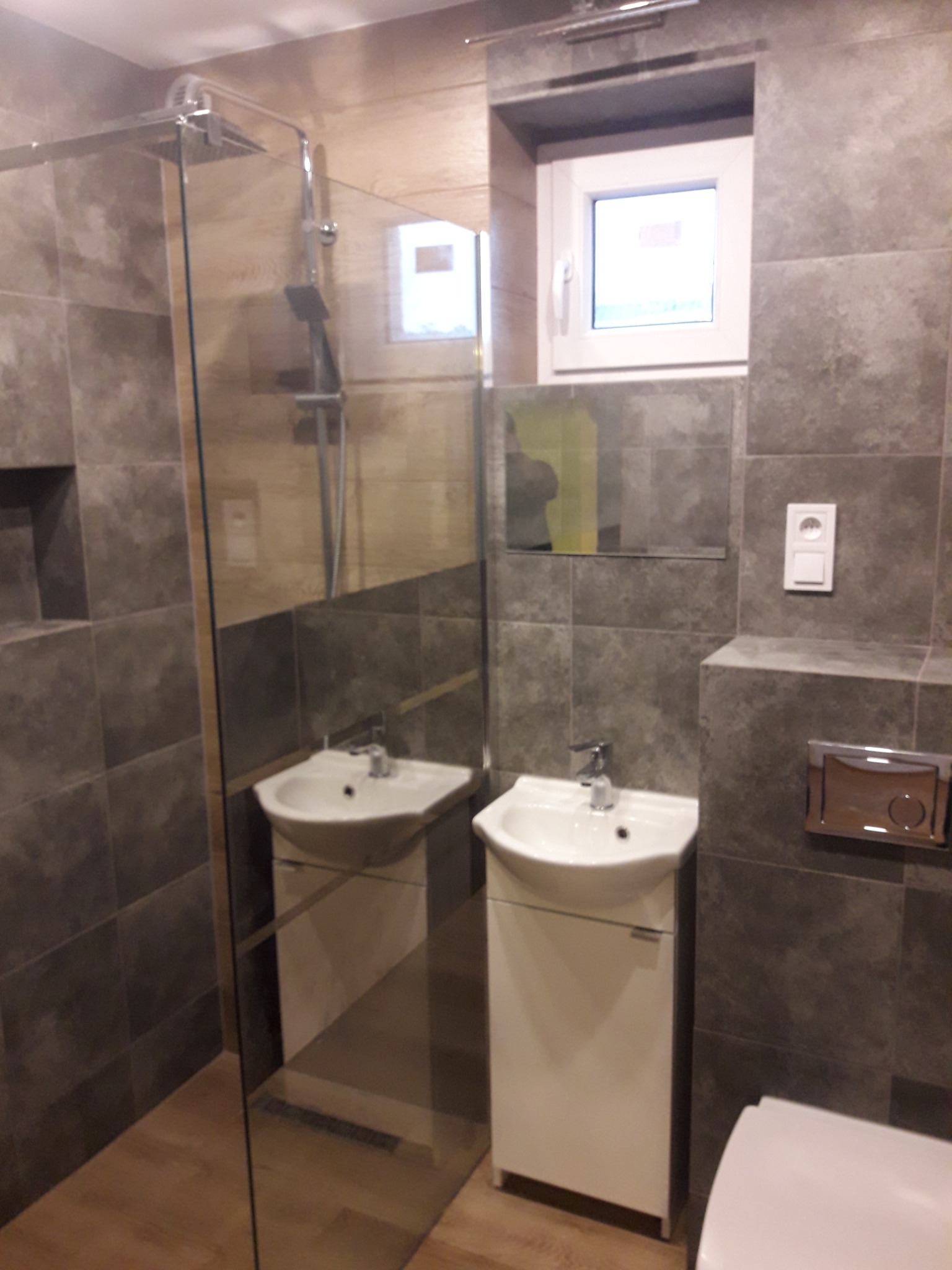 Szara łazienka z kabiną prysznicową typu walk-in, umywalką na szafce i wiszącą toaletą, oświetlona naturalnym światłem z okna.