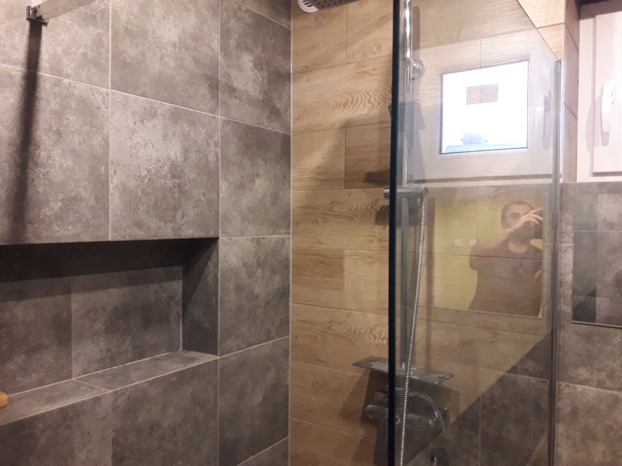 Nowoczesna kabina prysznicowa z szarymi płytkami imitującymi kamień i drewnopodobnymi płytkami na ścianie, widoczny wnęka w ścianie wyłożona szarymi płytkami, szklane drzwi kabiny, w odbiciu osoba...