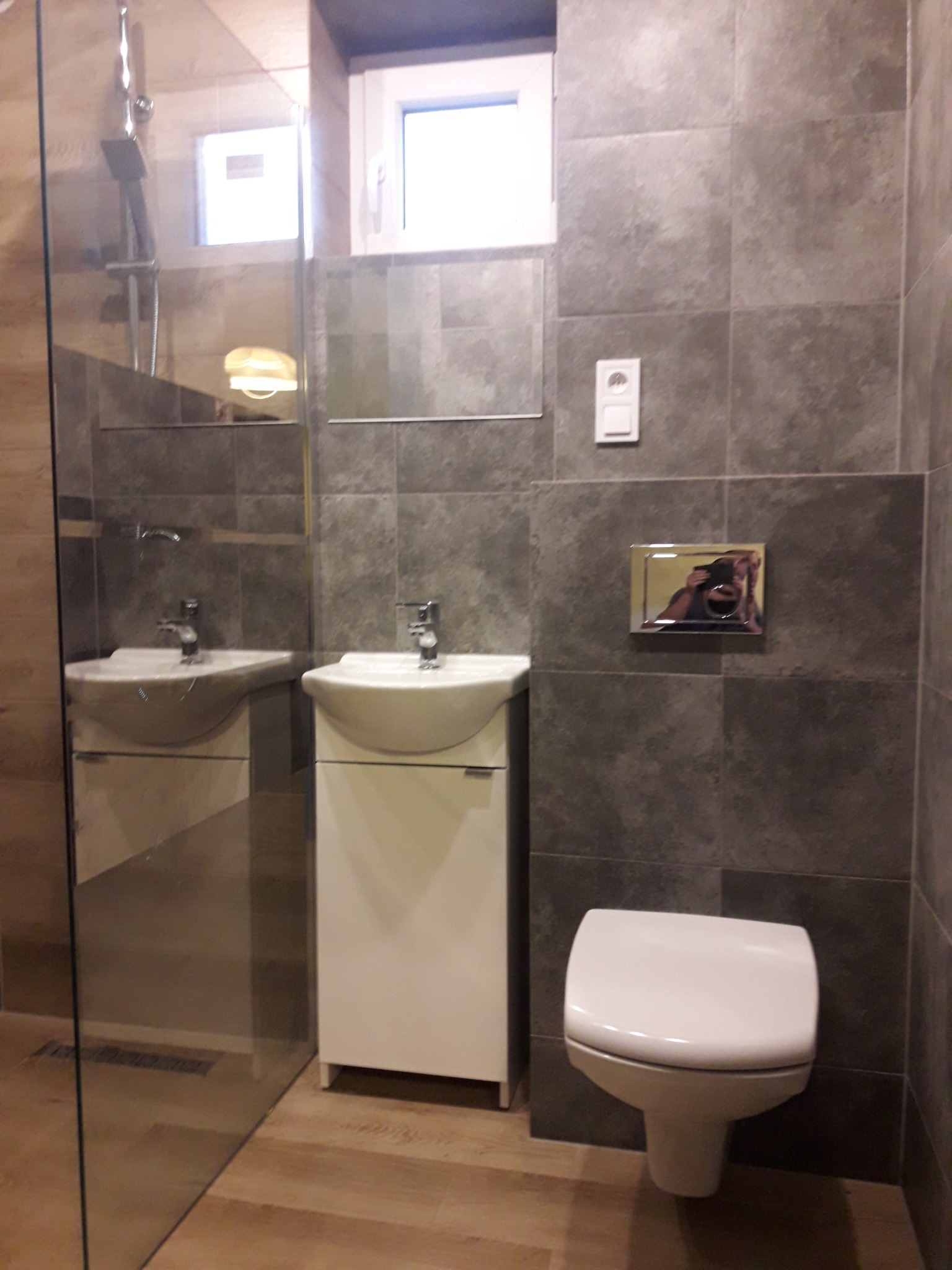 Nowoczesna łazienka z szarymi płytkami, kabiną prysznicową ze szkła, białą umywalką z szafką i podwieszaną toaletą. W tle widoczne okno i odbicie osoby robiącej zdjęcie w przycisku spłuczki.