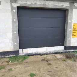 FC SPÓŁKA Z OGRANICZONĄ ODPOWIEDZIALNOŚCIĄ - Zewnętrzna, segmentowa brama garażowa w kolorze grafitowym zamontowana w surowym stanie budowy, widoczna tablica informacyjna budowy po prawej stronie.