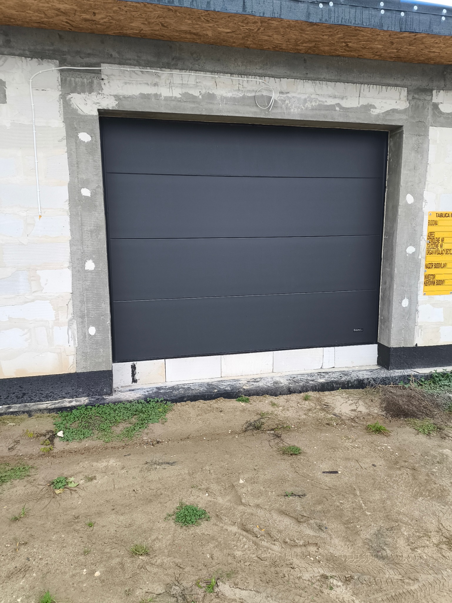 Zewnętrzna, segmentowa brama garażowa w kolorze grafitowym zamontowana w surowym stanie budowy, widoczna tablica informacyjna budowy po prawej stronie.