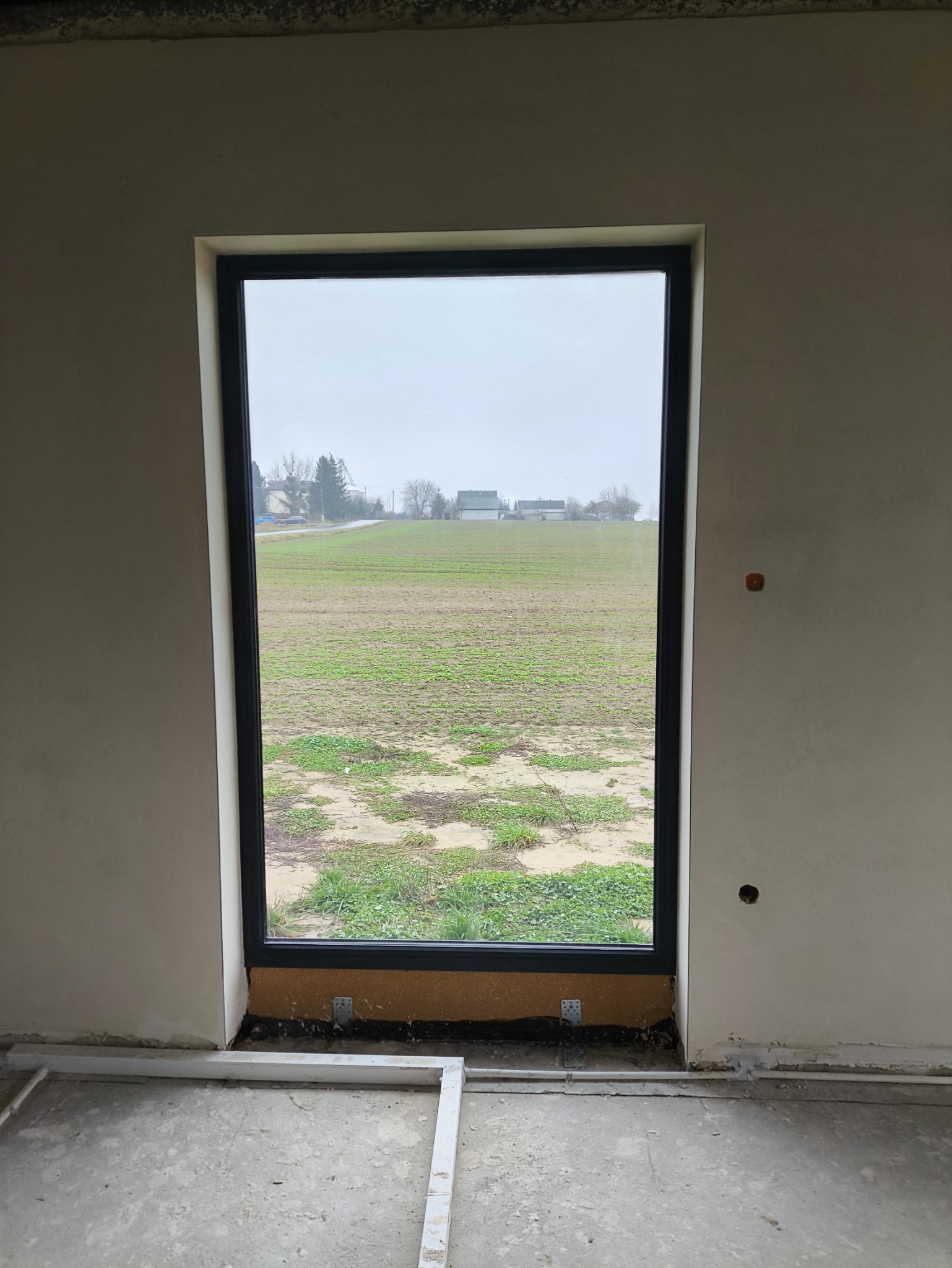 Nowe okno PCV z czarną ramą, zamontowane w surowym wnętrzu z widokiem na pole uprawne i zabudowania w oddali.