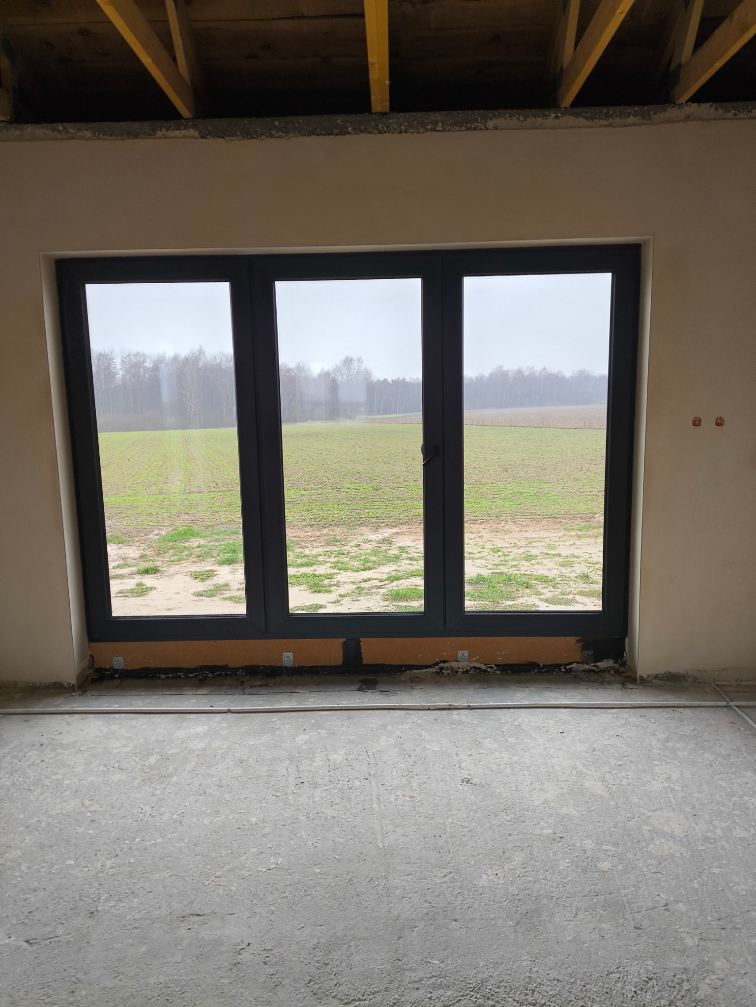 Trzyszybowe okno PCV w kolorze antracytowym, zamontowane w surowym stanie, z widokiem na pole i las we mgle.
