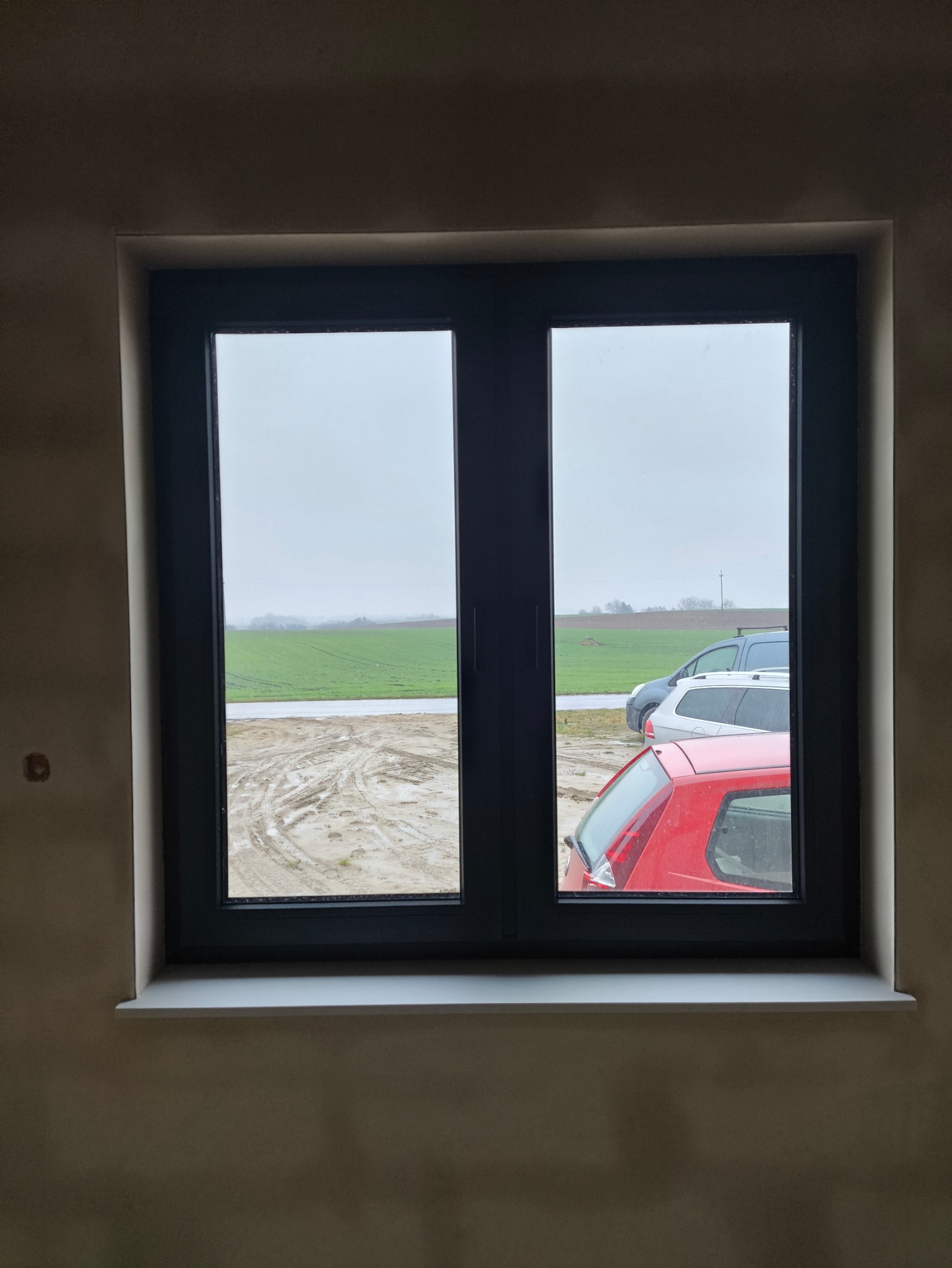 Zamknięte, dwuskrzydłowe okno PCV w ciemnej ramie z widokiem na pole, błotniste koleiny i zaparkowane samochody w pochmurny dzień.
