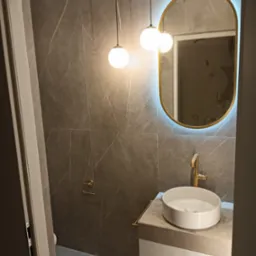 Nowoczesna łazienka z szarymi płytkami imitującymi marmur, okrągłą umywalką nablatową i podświetlanym lustrem w złotej ramie. Minimalistyczne oświetlenie.