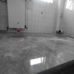 Bartko remonty i wykończenia - Wykończona łazienka z szarymi, błyszczącymi płytkami podłogowymi. Na ścianach białe, kwadratowe płytki. Widoczny grzejnik i butelka. Minimalistyczny design.