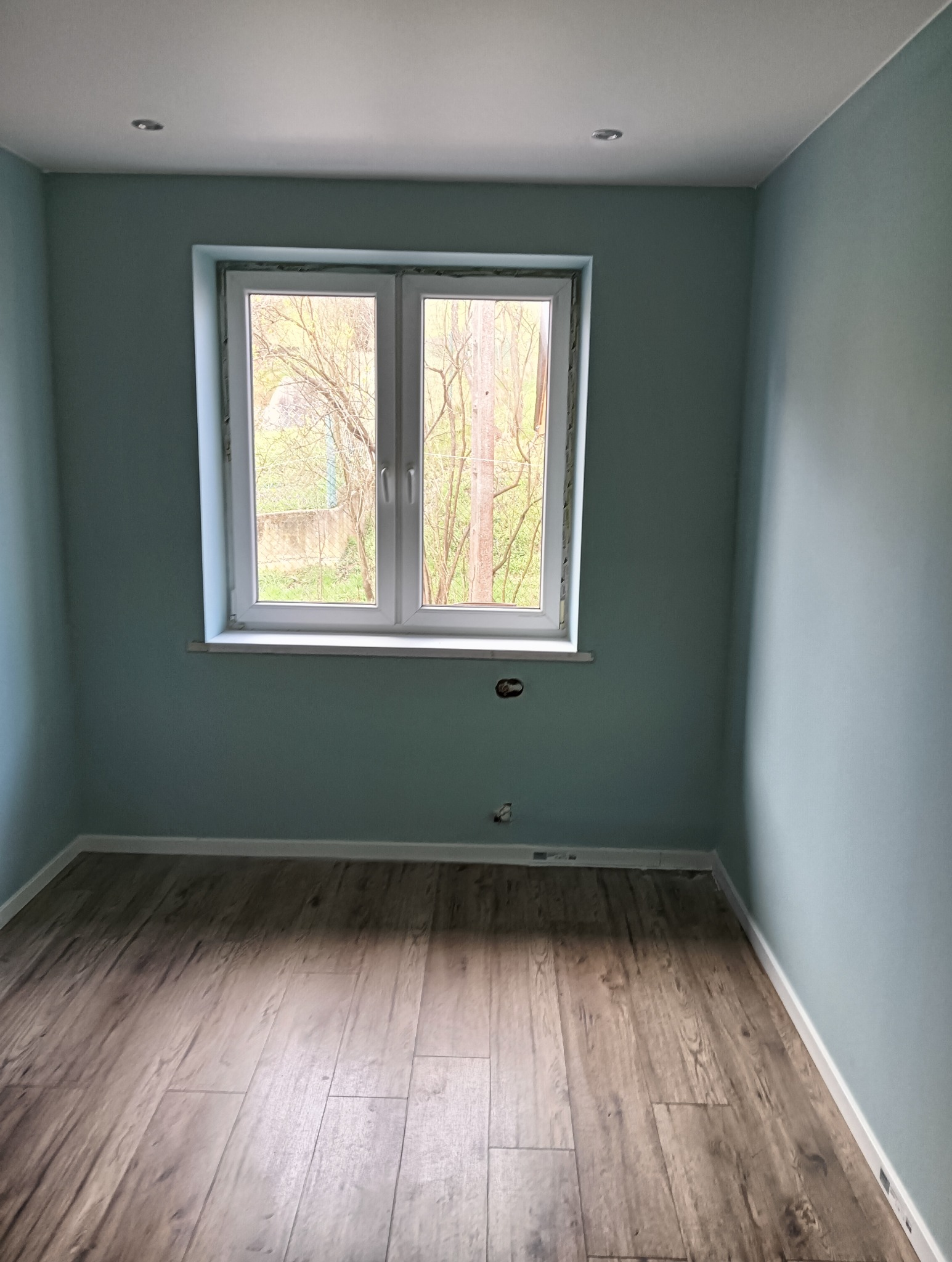Pokój po remoncie: panele podłogowe w odcieniach brązu, błękitne ściany, białe okno z widokiem na drzewa. Widoczne gniazdka elektryczne.